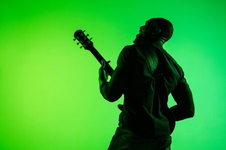 Young african-american musician playing the guitar like a rockstar on gradient green-yellow background. Concept of music, hobby, festival, open-air. Joyful attractive guy improvising, singing song.の写真素材