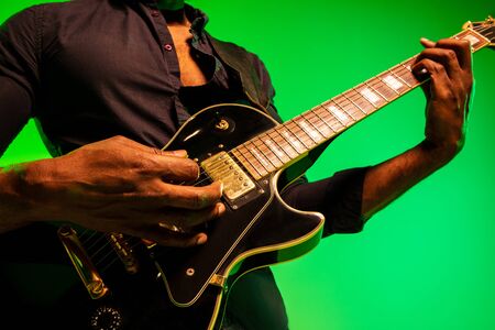 Young african-american musician playing the guitar like a rockstar on gradient green-yellow background. Concept of music, hobby, festival, open-air. Joyful attractive guy improvising, singing song.の写真素材