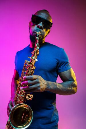 Young african-american jazz musician playing the saxophone on pink studio background in trendy neon light. Concept of music, hobby. Joyful attractive guy improvising. Colorful portrait of artist.の写真素材