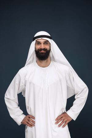 Half-length portrait of arabian saudi businessman on dark blue studio background. Young male model standing and smiling. Concept of business, finance, facial expression, human emotions.の写真素材