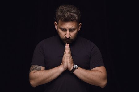 Close up portrait of young man isolated on black studio background. Photoshot of real emotions of male model. Praying with closed eyes, looks hopeful. Facial expression, human emotions concept.の写真素材