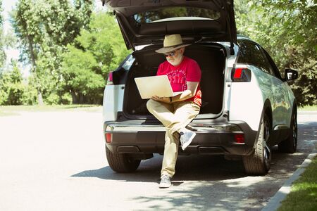 Senior man sitting on his cars trunk and working outdoors at the citys street in sunny day. Male model in sunglasses using notebook. Concept of working, business, job, start-up, freelance.の写真素材