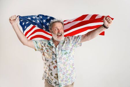 Celebrating an Independence day. Stars and Stripes. Senior man with the flag of the United States of America isolated on white studio background. Looks crazy happy and proud as a patriot of his country.の写真素材