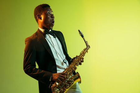 Young african-american jazz musician playing the saxophone on gradient yellow-green studio background. Concept of music, hobby, festival. Joyful attractive guy improvising. Colorful portrait of artist.の写真素材