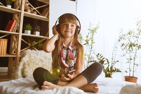Girl using different gadgets at home. Little model sitting in her bed with big headphones listening to favourite music and enjoying. Concept of interaction of kids and modern technologies.の写真素材