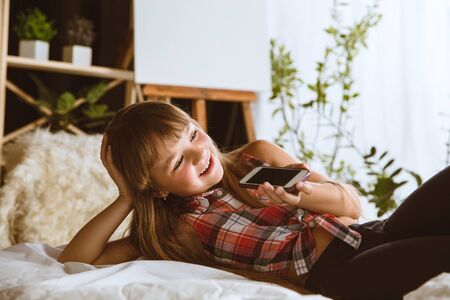 Girl using different gadgets at home. Little model lying in her bed with smartphone and talking with her friends in voices massages or live chats. Concept of interaction of kids and modern technologies.の写真素材