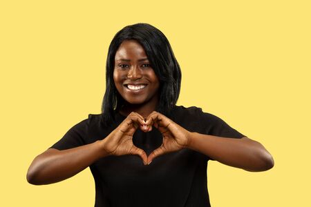 Young african-american woman isolated on yellow studio background, facial expression. Beautiful female half-length portrait. Concept of human emotions, facial expression. Showing the sign of heart.の写真素材