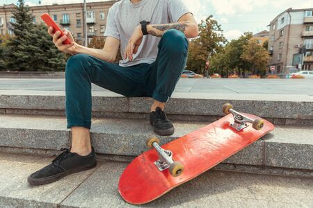 Skateboarder resting after riding at the citys street in sunny day. Young man in sneakers and cap with a longboard on the asphalt. Concept of leisure activity, sport, extreme, hobby and motion.の写真素材