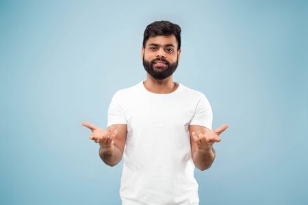 Half-length close up portrait of young hindoo man in white shirt on blue background. Human emotions,...