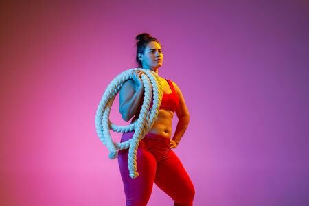 Young caucasian plus size female models doing exercises on gradient purple background in neon light. Cross training with the rope. Concept of sport, healthy lifestyle, body positive, equality.の写真素材