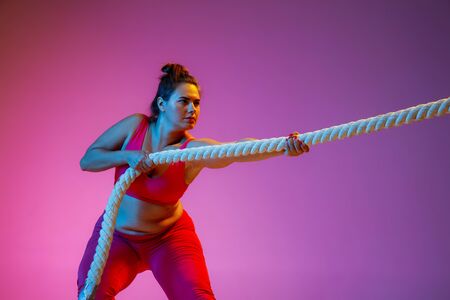 Young caucasian plus size female models doing exercises on gradient purple background in neon light....