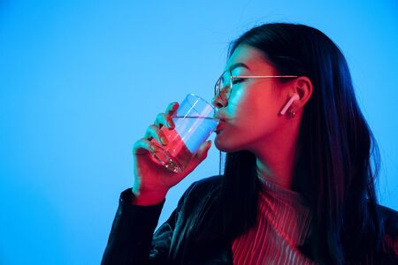 Beautiful female half-length portrait isolated on blue studio background in neon light. Young emotional woman. Human emotions, facial expression concept. Drinking water, listen to music wirelessly.の写真素材