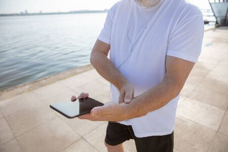 Senior man as runner with fitness tracker at the riverside. Caucasian male model using gadgets while...