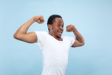 Half-length close up portrait of young african-american man in white shirt on blue background. Human emotions, facial expression, ad, concept. Celebrating, wondered, astonished, shocked, crazy happy.の写真素材