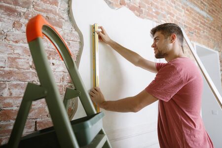 Young man preparing for doing apartment repair by hisselfes. Before home makeover or renovation. Concept of relations, family, DIY. Measuring the wall before painting or design making.の写真素材