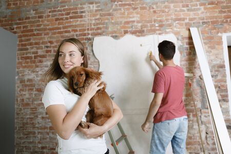Young couple doing apartment repair together themselves. Married man and woman doing home makeover or renovation. Concept of relations, family, pet, love. Painting the wall, holding the dog.の写真素材