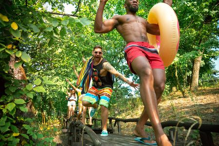 Group of happy friends having fun while running to swim on river. Joyful women and men in swimsuit...