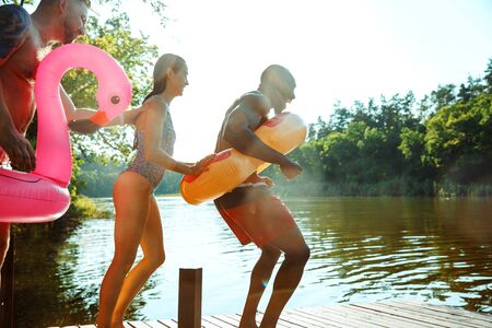 Happy friends having fun while laughting and jumping from the pier in river. Joyful male and female...