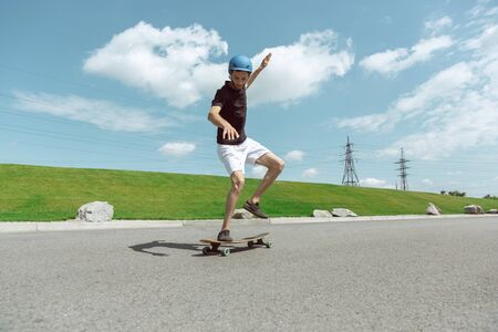 Skateboarder doing a trick at the citys street in sunny day. Young man in equipment riding and longboarding near by meadow in action. Concept of leisure activity, sport, extreme, hobby and motion.の写真素材