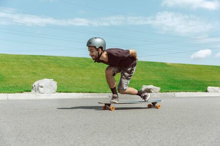 Skateboarder doing a trick at the citys street in sunny day. Young man in equipment riding and longboarding near by meadow in action. Concept of leisure activity, sport, extreme, hobby and motion.の写真素材