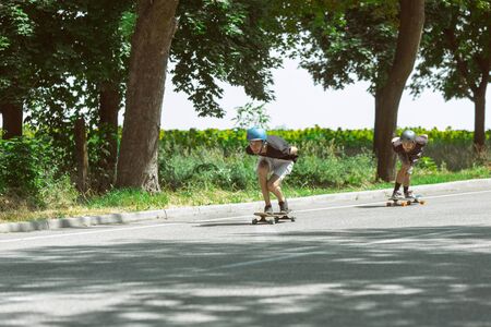 Skateboarders doing a trick at the citys street in sunny day. Young men in equipment riding and longboarding near by meadow in action. Concept of leisure activity, sport, extreme, hobby and motion.の写真素材