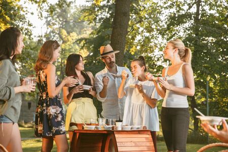Group of happy friends eating and drinking beers at barbecue dinner on sunset time. Having meal together outdoor in a forest glade. Celebrating and relaxing. Summer lifestyle, food, friendship concept.の写真素材