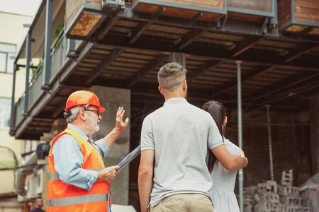 Foreman or achitect engineer shows future house, office or store design plans to a young couple. Meeting at the construction site to talk about facade appearance, interior decoration, home layout.の写真素材