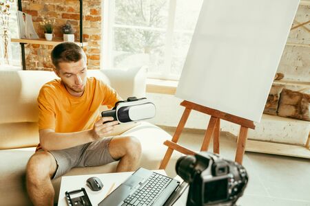 Young caucasian male blogger with professional equipment recording video review of VR glasses at home. Blogging, videoblog, vlogging. Man evaluates virtual reality headset while streaming live.の写真素材