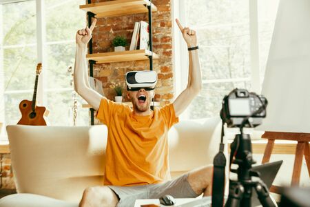 Full of emotions. Caucasian male blogger with professional camera recording video review of VR glasses at home. Blogging, videoblog, vlogging. Man using virtual reality headset while streaming live.の写真素材
