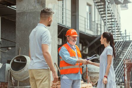 Foreman or achitect engineer shows future house, office or store design plans to a young couple. Meeting at the construction site to talk about facade appearance, interior decoration, home layout.の写真素材