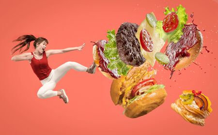 Fast food. Young caucasian woman fights with unhealthy nutrition. Kicks burger or sandwich in jump on red background. Copyspace for your ad. Creative collage about food, weight loss, healthy eating.の写真素材