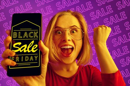 Portrait of young caucasian woman showing mobile phone screen on purple background with neon...