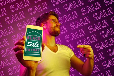 Portrait of young caucasian man showing mobile phone screen on purple background with neon lettering. Concept of sales, black friday, cyber monday, finance, business. Online shops and payments bill.の写真素材