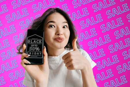Portrait of young caucasian woman showing mobile phone screen on pink background with neon lettering. Concept of sales, black friday, cyber monday, finance, business. Online shops and payments bill.の写真素材