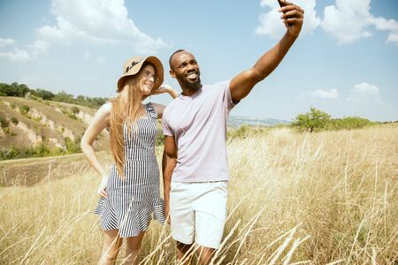 Young multiethnic international couple outdoors at the meadow in sunny summer day. African-american man and caucasian woman having picnic together. Concept of relationship, summertime. Making selfie.の写真素材