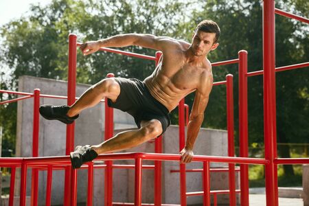 Young muscular shirtless caucasian man jumping above horizontal bar at playground in sunny summers...