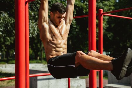 Young muscular shirtless caucasian man doing pull-ups on horizontal bar at playground in sunny...