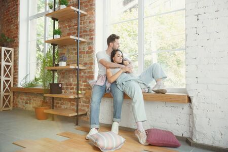 Lets catch this moment. Young couple moved to a new house or apartment. Look happy and confident. Family, moving, relations, first home concept. Sitting near by window, hugging and making selfie.の写真素材
