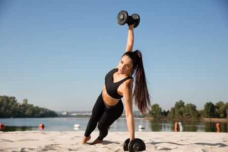 Young healthy woman training upper body with weights at the beach. Single caucasian female model...