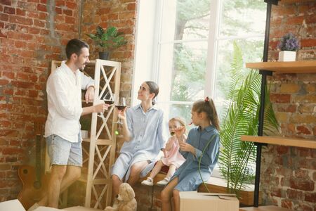 New life. Adult family moved to a new house or apartment. Spouses and children look happy and confident. Moving, relations, new life concept. Playing together, sitting near by window, drinking wine.の写真素材