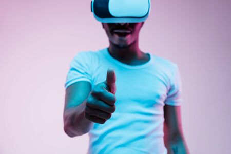 Young african-american man in VR-glasses in neon light on gradient background. Male portrait. Concept of human emotions, facial expression, modern gadgets and technologies. Pointing empty search bar.の写真素材