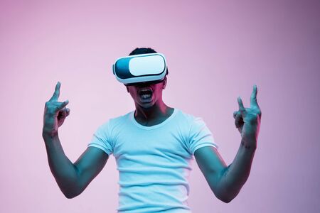 Young african-american mans playing in VR-glasses in neon light on gradient background. Male portrait. Concept of human emotions, facial expression, modern gadgets and technologies. Looks like winner.の写真素材