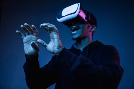 Young african-american man in VR-glasses in neon light on blue background. Male portrait. Concept of...