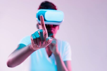 Portrait of young african-american womans playing in VR-glasses in neon light on gradient background. Concept of human emotions, facial expression, modern gadgets and technologies. Touches empty bar.の写真素材