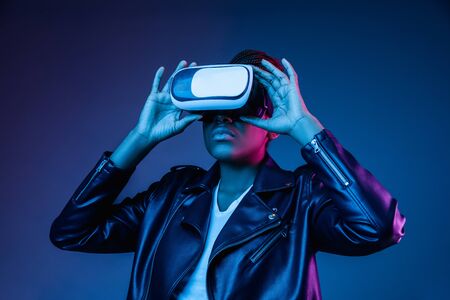 Portrait of young african-american womans playing in VR-glasses in neon light on blue background. Concept of human emotions, facial expression, modern gadgets and technologies. Look confident.の写真素材