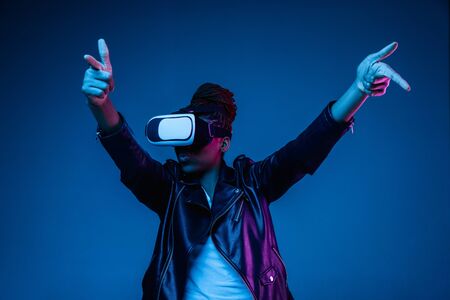 Portrait of young african-american womans playing in VR-glasses in neon light on blue background. Concept of human emotions, facial expression, modern gadgets and technologies. Look shooting, pointing.の写真素材