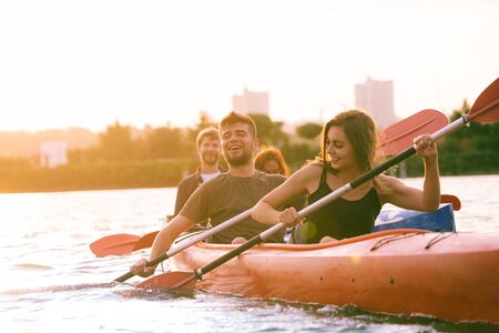 Happy young caucasian group of friends kayaking on river with sunset in the backgrounds. Having fun in leisure activity. Happy male and female model laughting on the kayak. Sport, relations concept.の写真素材