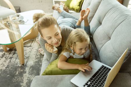 Female caucasian teacher and little girl, or mom and daughter. Homeschooling. Lying on the sofa and using laptop for knowledges getting while lesson is. Education, school, studying concept.の写真素材