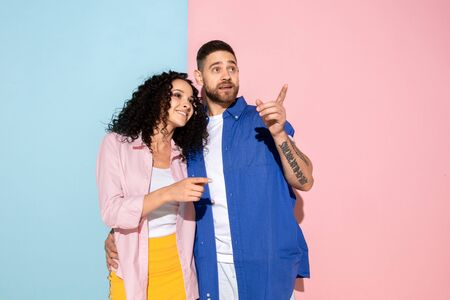 Young emotional caucasian couple in bright casual clothes posing on pink and blue background....