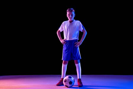 Young boy as a soccer or football player in sportwear standing with the ball on dark studio background. Fit playing boy in action, movement, motion at game. Purple neon light. Look confident.の写真素材